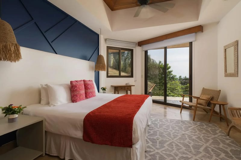 Casa Anjali Luxury Villa · 34. This bedroom features a king bed, a dark blue geometric accent wall, a vaulted teak ceiling with a fan, and floor-to-ceiling glass doors providing rainforest and distant ocean views from a private terrace.