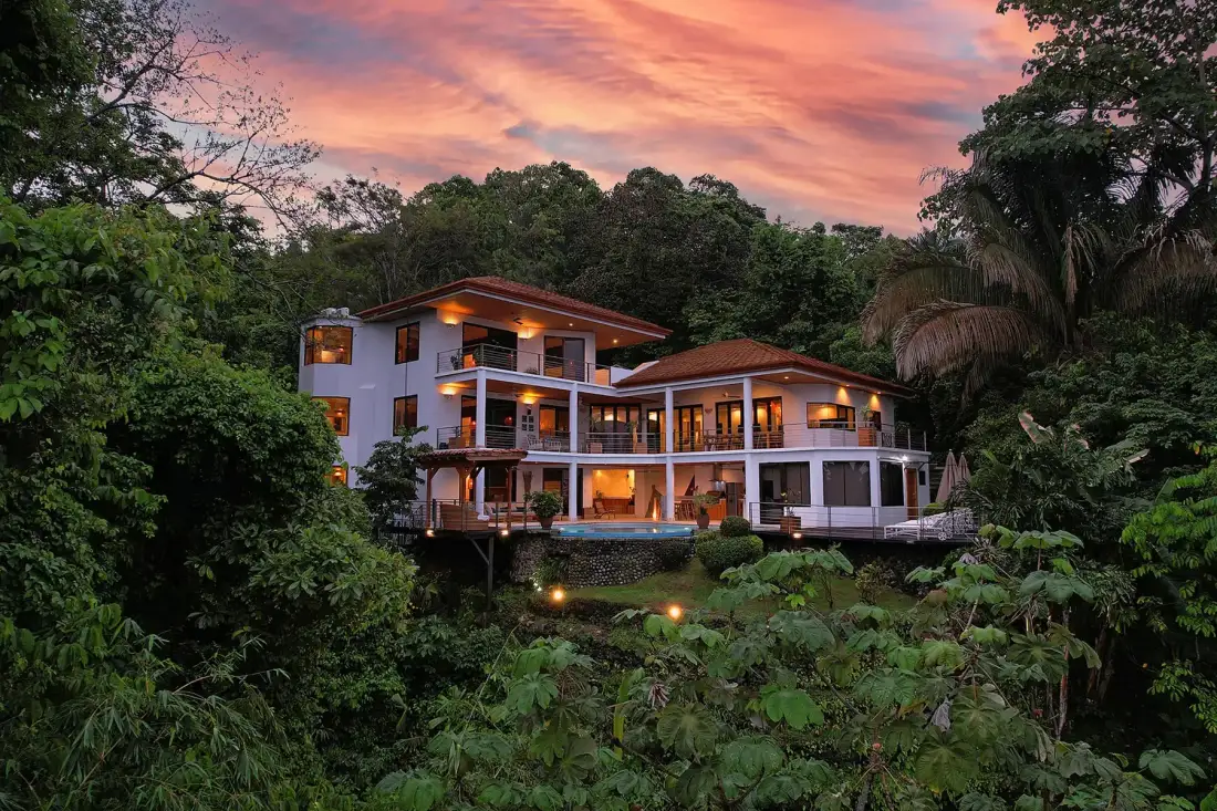 A multi-level house glows among dense greenery beneath a colorful evening sky.