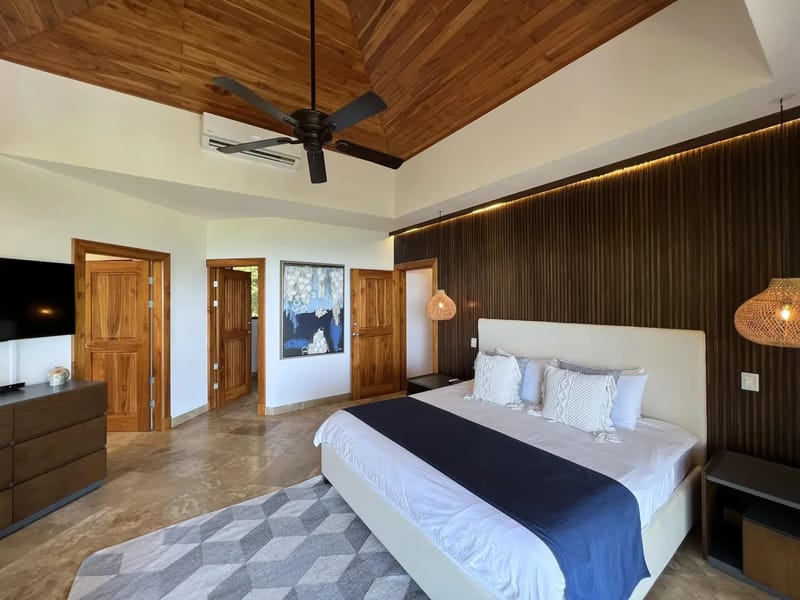 Casa Anjali Luxury Villa · 25. This king bedroom features a vaulted teak ceiling, a dark wood accent wall with integrated lighting, tiled floors, and wooden doors opening to natural light and views of the surrounding rainforest.