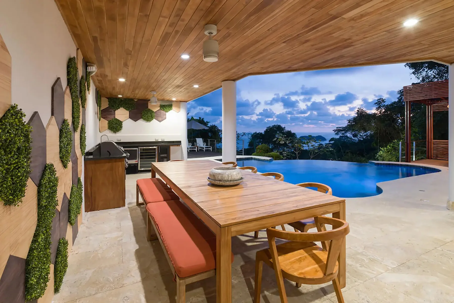 A covered outdoor dining area overlooks a swimming pool and tree-covered landscape.