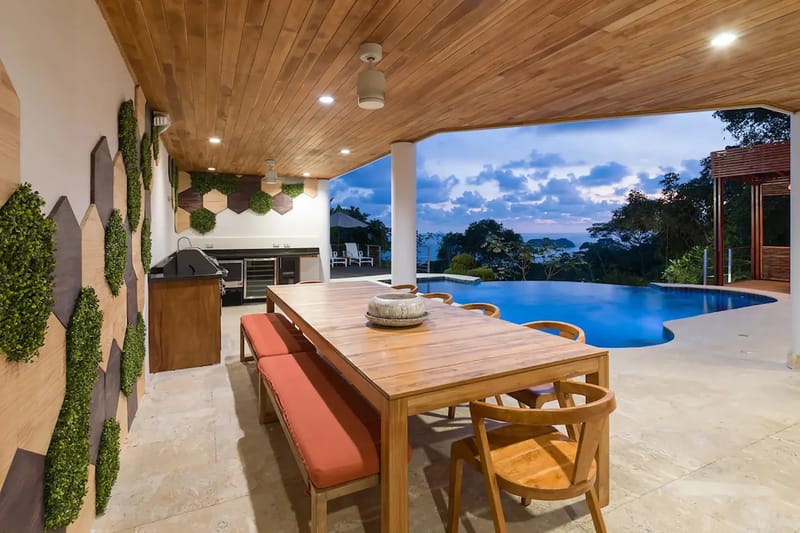 Casa Anjali Luxury Villa · 17. This open-air, covered terrace features a wooden dining table and an integrated outdoor kitchen, all overlooking an infinity pool and the ocean framed by a twilight rainforest.