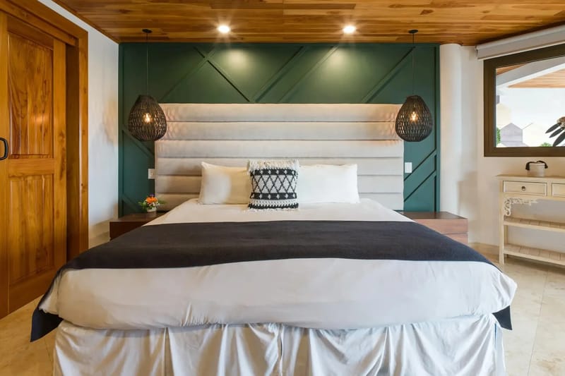 Casa Anjali Luxury Villa · 70. A king bedroom features a rich wood-paneled ceiling, a green geometric accent wall, and natural light entering from a window overlooking the surroundings.