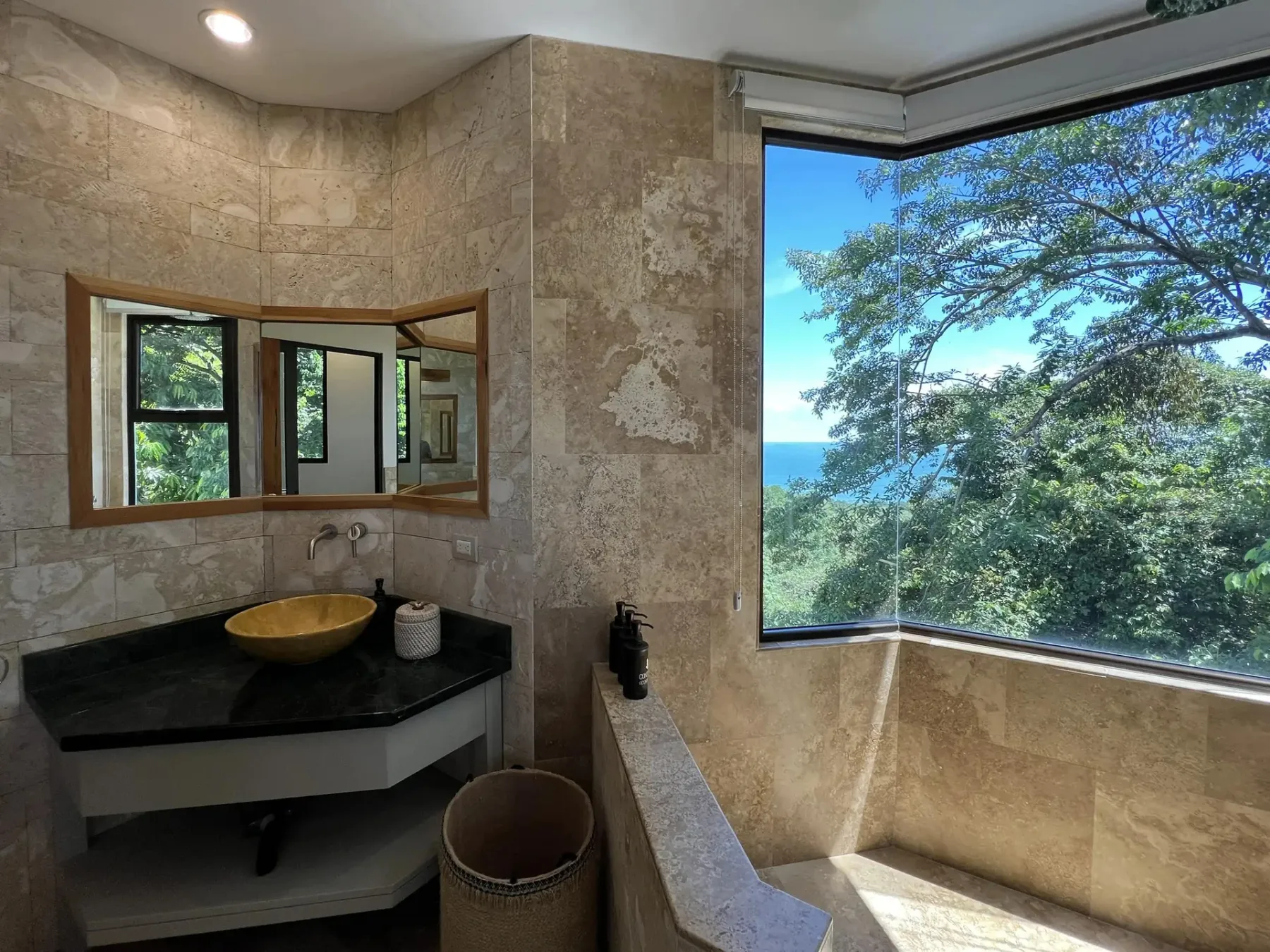 A tiled bathroom with a sink and a large window overlooking trees and the ocean.