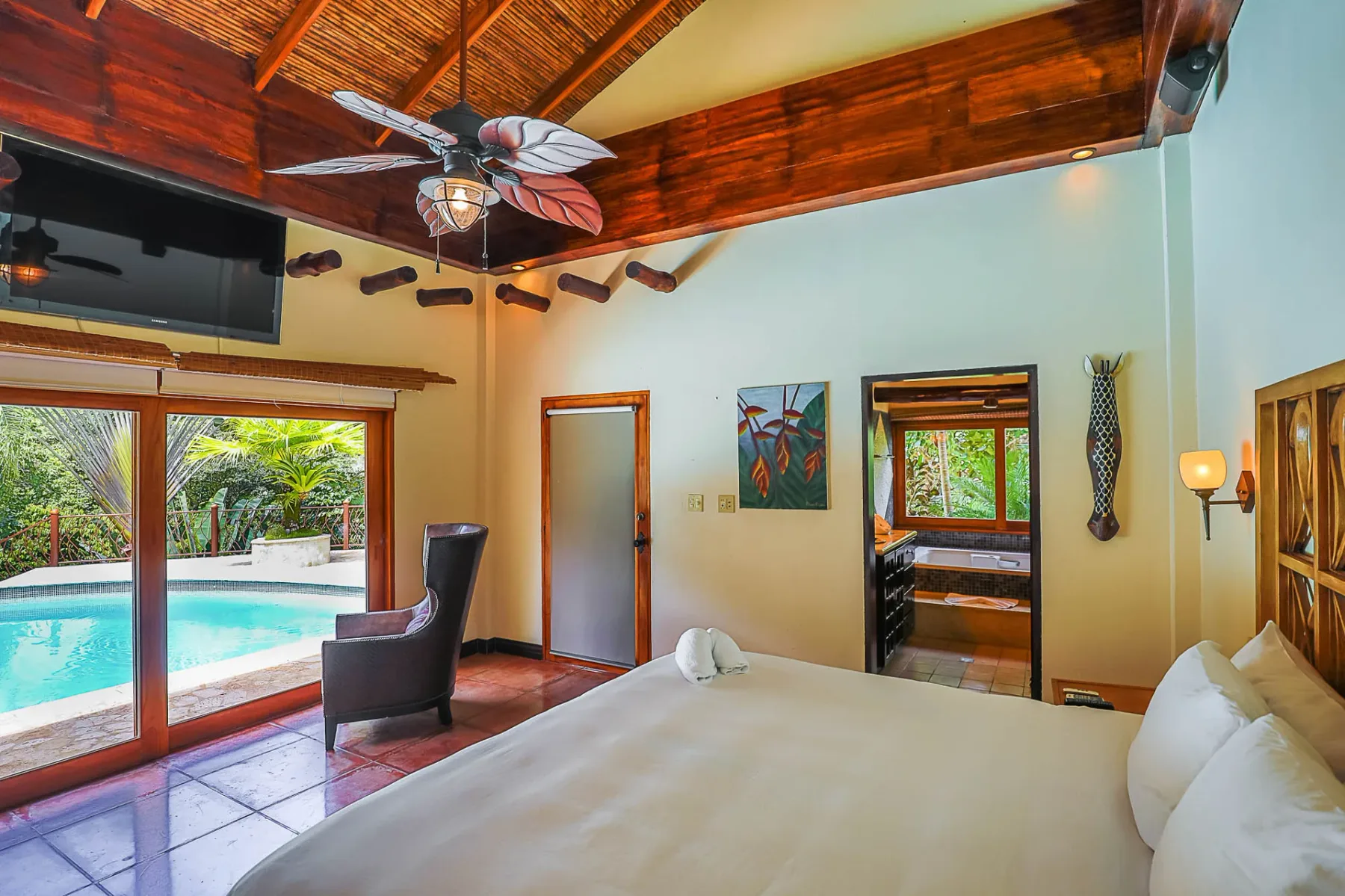 A bedroom with a large bed, exposed wooden ceiling beams, and sliding glass doors overlooking a pool.