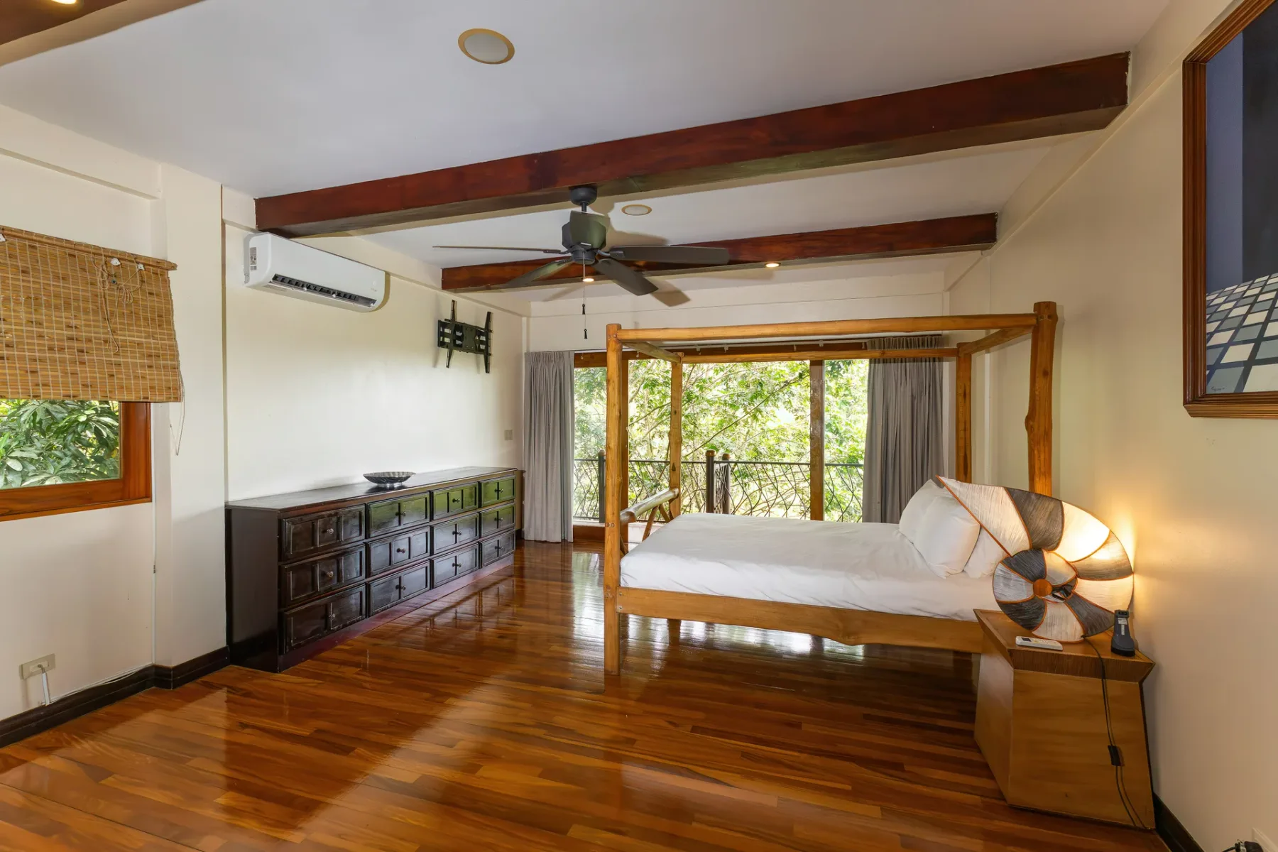 A bedroom with a wooden bed, dark dresser, and windows overlooking greenery.