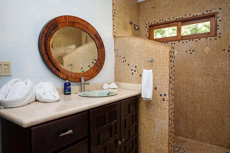 Casa Estrella del Mar · 48. The bathroom features a dark wood vanity with a vessel sink and a large wood-framed mirror, complemented by a mosaic-tiled shower with a window that provides natural light and views of tropical greenery.