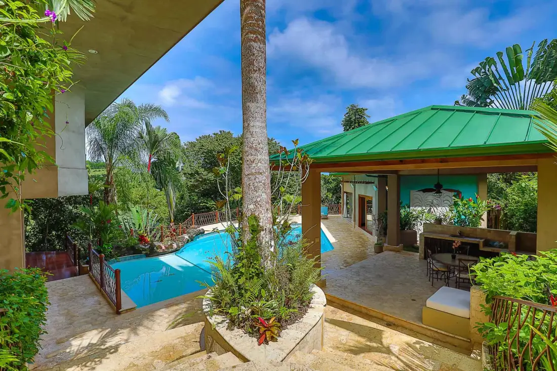 An outdoor pool beside a covered pavilion, surrounded by tropical greenery.