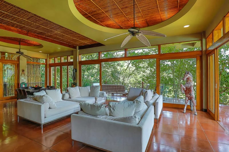 Casa Estrella del Mar · 20. An open living area with comfortable white sofas and a bamboo ceiling is illuminated by natural light through expansive windows offering direct views of the surrounding rainforest.