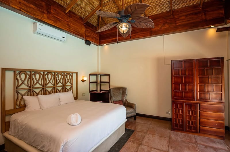 Casa Estrella del Mar · 35. This bedroom features a high vaulted ceiling with exposed wood beams and natural woven panels, a large bed with a decorative wooden headboard, a spacious wooden wardrobe, and a tropical leaf-bladed ceiling fan, offering a comfortable and rustic ambiance.