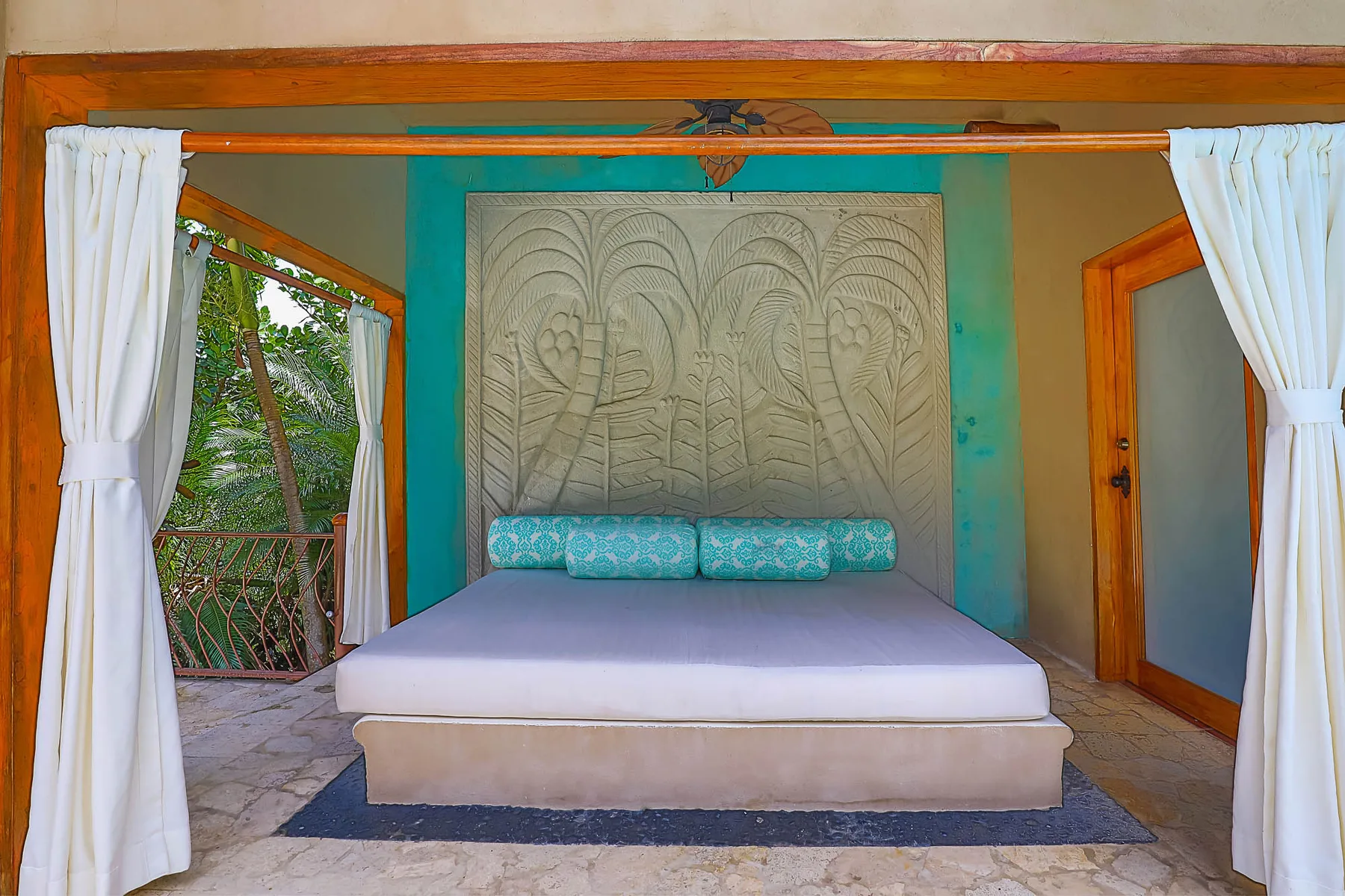 A bedroom with a low bed beneath a carved wall panel and white curtains.
