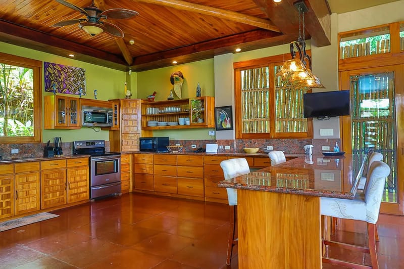 Casa Estrella del Mar · 9. This kitchen, featuring extensive wooden cabinetry, granite countertops, and a large island, provides tropical views through wooden-framed windows and is capped by a high wood-paneled ceiling, offering a warm and open feel.