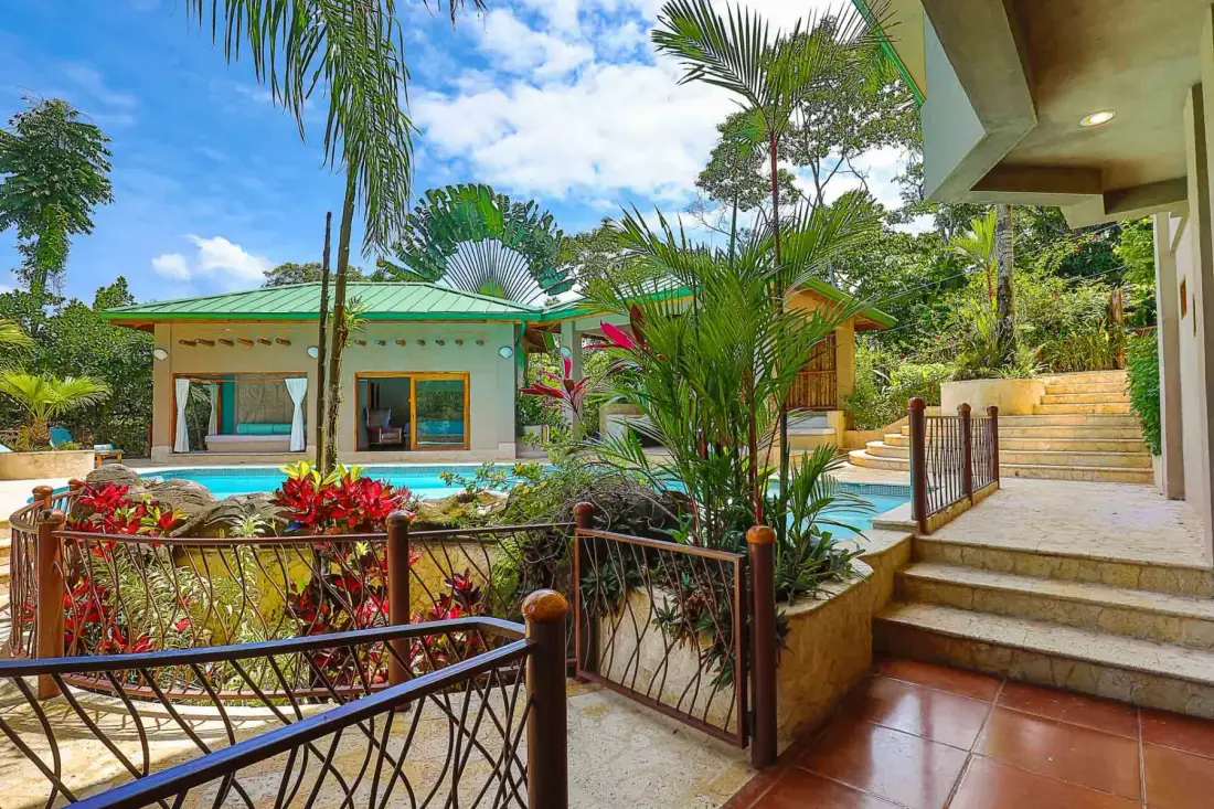 An outdoor pool sits among tropical plants, with a gated walkway and steps beside it.