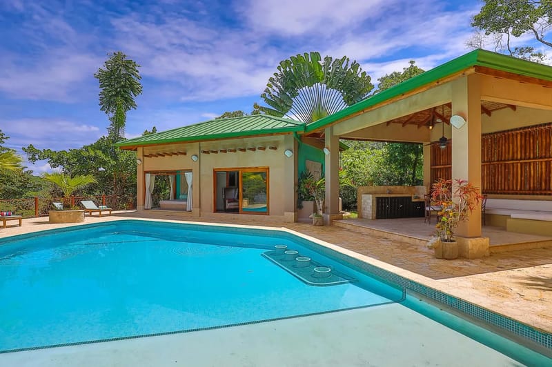 Casa Estrella del Mar · 16. A bright blue swimming pool fronts a pool house with a visible bedroom and an open-air pavilion featuring an outdoor kitchen and lounge area, all surrounded by tropical greenery.