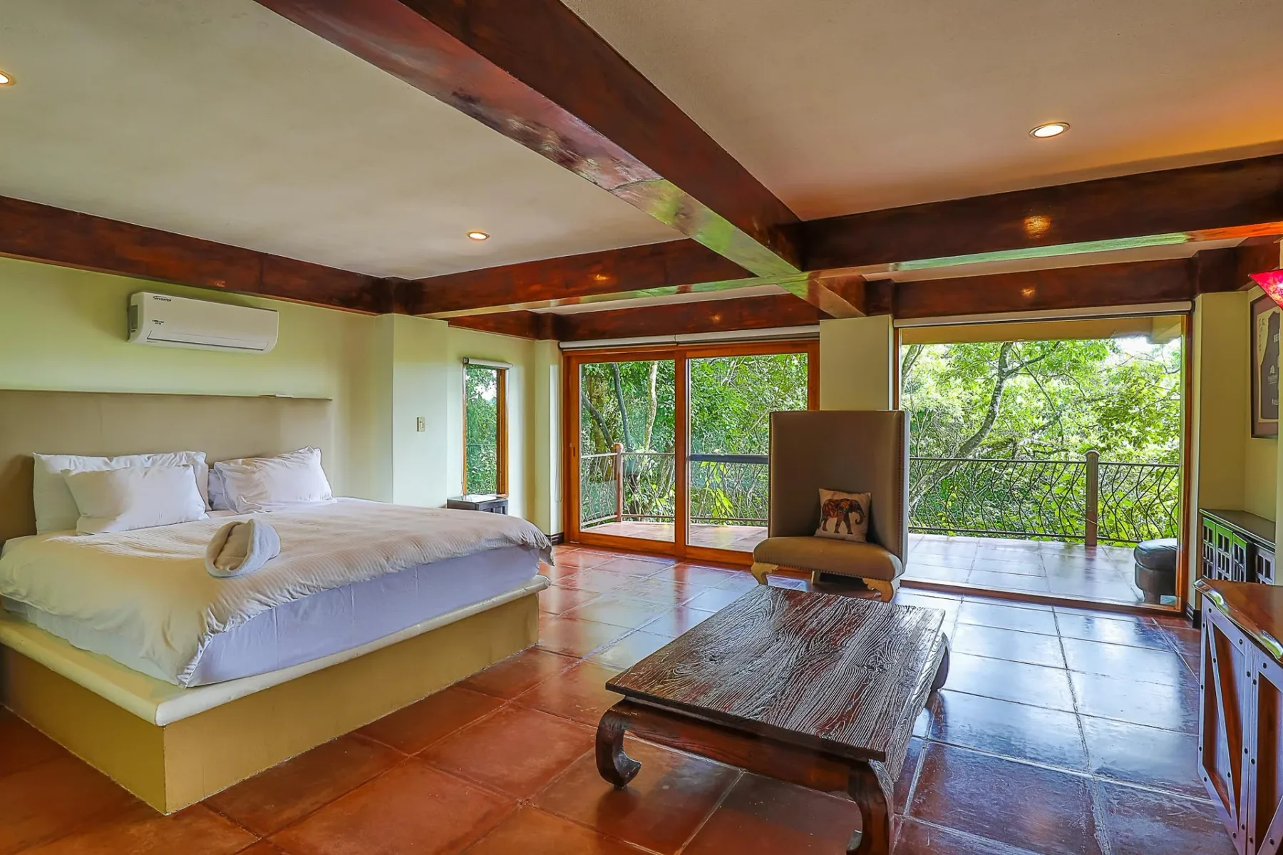 A bedroom with a large bed and broad windows overlooking dense green foliage.