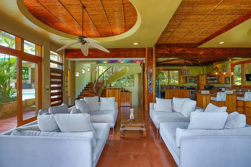 Casa Estrella del Mar · 21. An open-plan living room with comfortable white sofas features a high bamboo ceiling and direct access to a pool terrace, with a full kitchen and a spiral staircase visible within the tropical-view space.