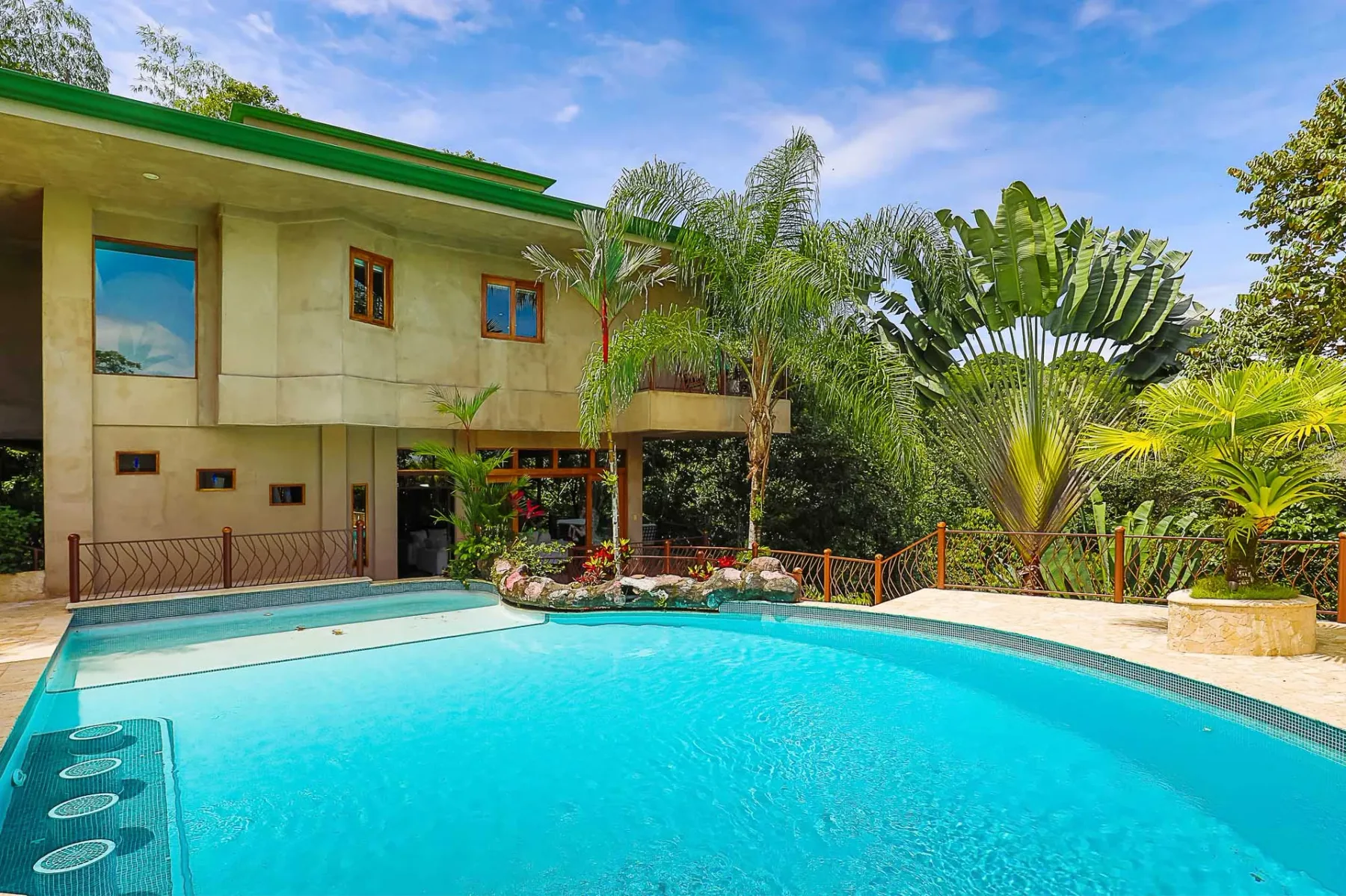 A modern house stands beside a swimming pool surrounded by lush tropical plants.