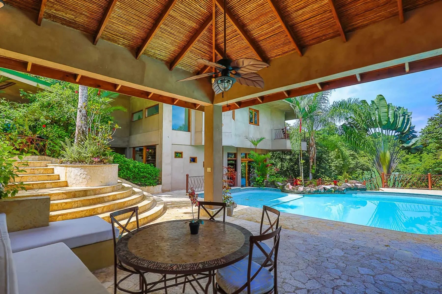 A covered poolside terrace has a round dining table overlooking the swimming pool and greenery.