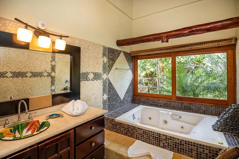 Casa Estrella del Mar · 46. A bathroom features a jetted tub positioned by a large window offering natural light and views of lush tropical foliage, complemented by a rustic wooden beam and decorative mosaic tile walls.