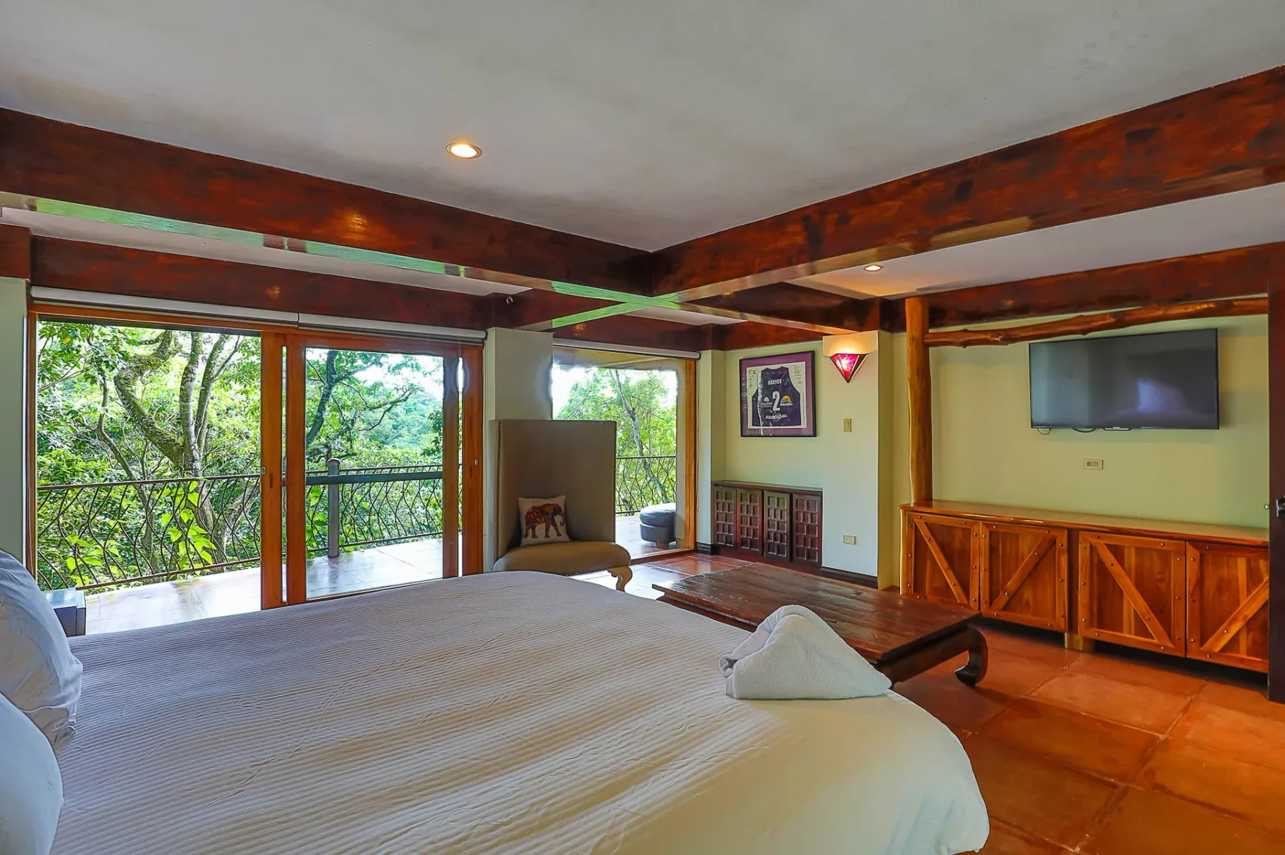 A bedroom with a large bed, wooden beams, and glass doors overlooking leafy trees.