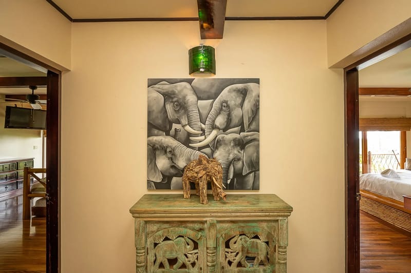 Casa Estrella del Mar · 27. A welcoming hallway featuring locally-sourced wood accents, a carved console, and elephant-themed artwork connects to a bright, natural-wood-furnished bedroom and a comfortable living area.