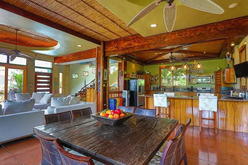 Casa Estrella del Mar · 25. This open-concept main floor combines a comfortable living area with sofas, a large wooden dining table, and a well-appointed kitchen, all featuring natural wood accents, ample light, and views of the lush rainforest.