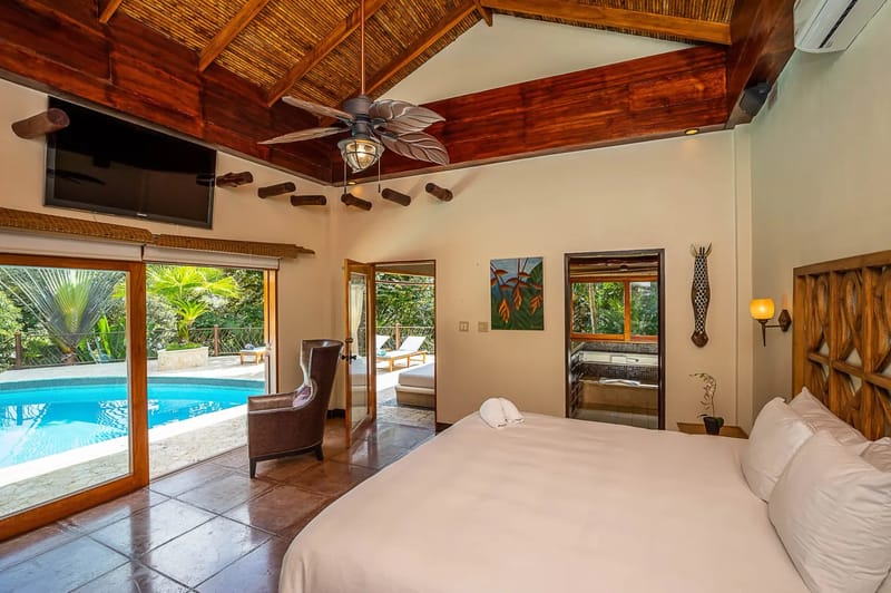 Casa Estrella del Mar · 43. A spacious bedroom features a vaulted ceiling with dark wood beams and bamboo, terracotta tile floors, direct access to a swimming pool, and an adjacent terrace, offering views of tropical foliage and ample natural light.