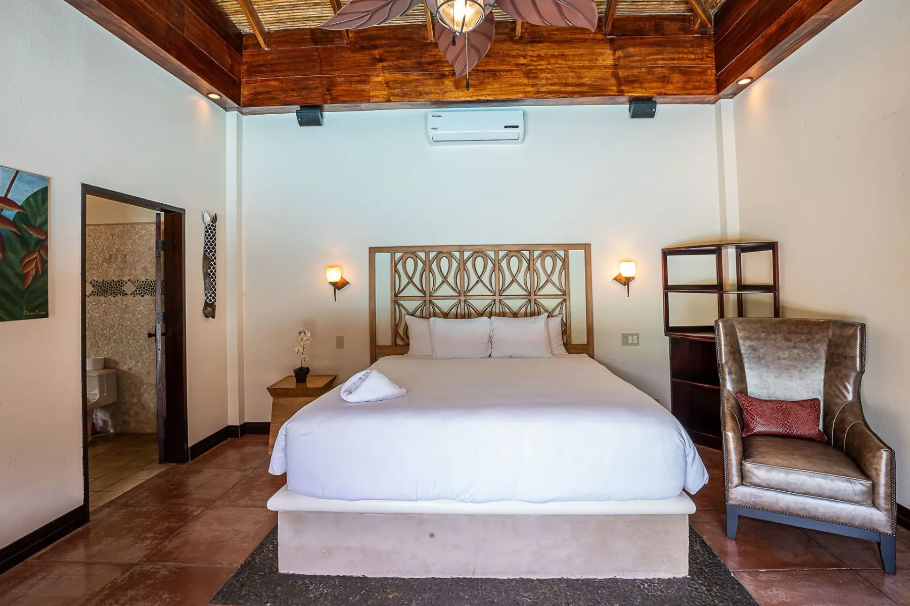 A bedroom with a bed beneath a high wooden ceiling and an air conditioner.