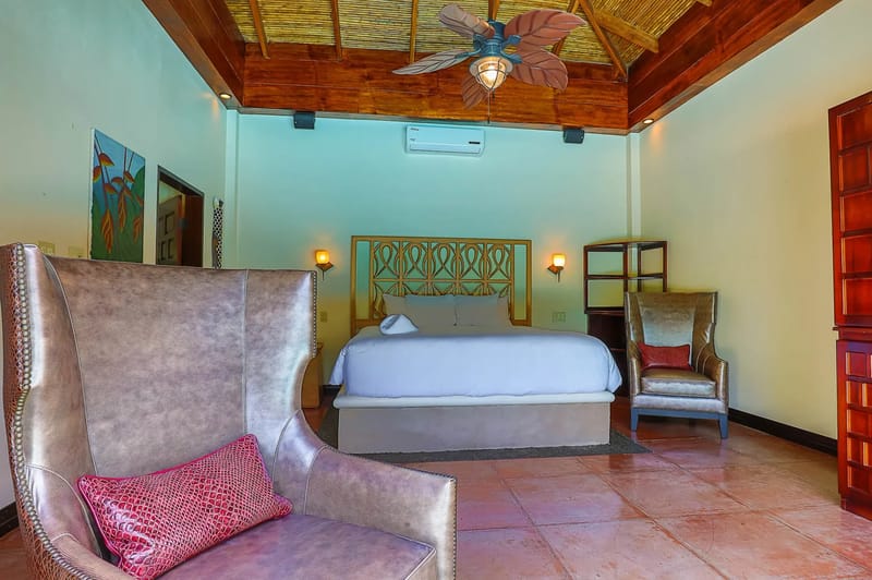Casa Estrella del Mar · 34. This inviting bedroom features a king bed with an ornate wooden headboard, a vaulted ceiling with exposed wooden beams and a tropical fan, and comfortable armchairs set on terracotta tile floors.