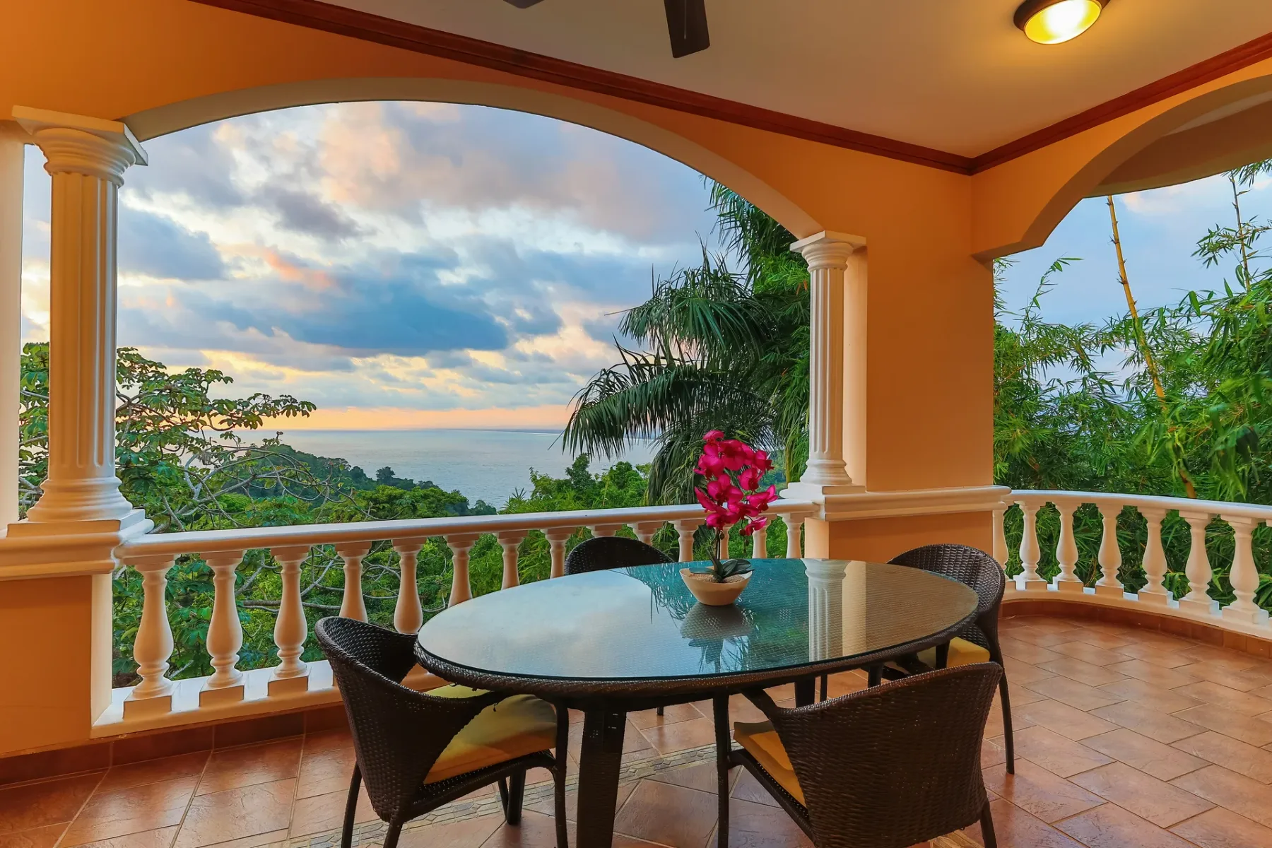 A covered terrace has a dining table overlooking the ocean and tropical greenery.