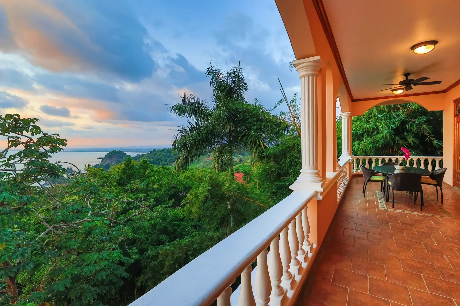 A covered balcony overlooks lush greenery and the ocean beneath a colorful evening sky.