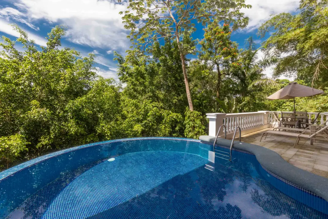 An outdoor swimming pool beside a patio with seating and surrounding trees.