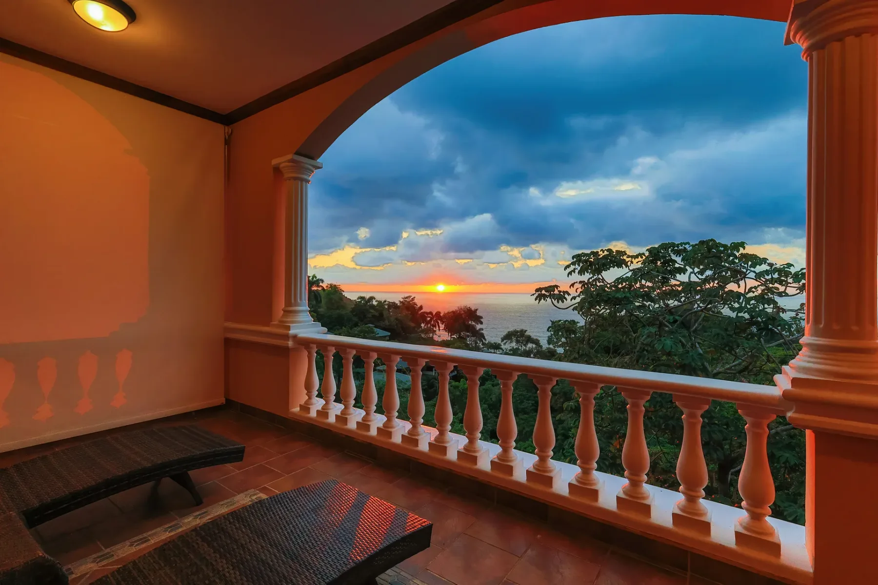 A covered balcony overlooks the ocean and trees at sunset.