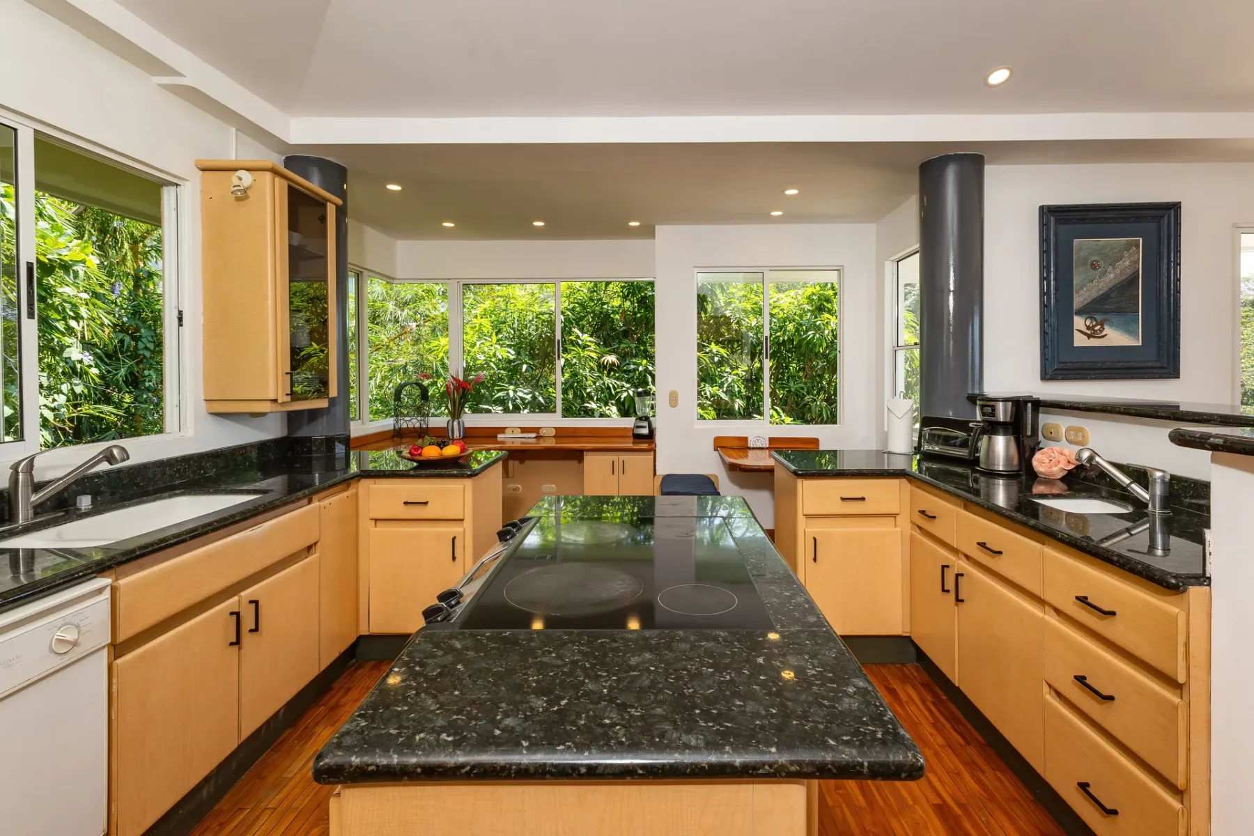 A bright kitchen with wooden cabinets, dark countertops, and windows overlooking greenery.