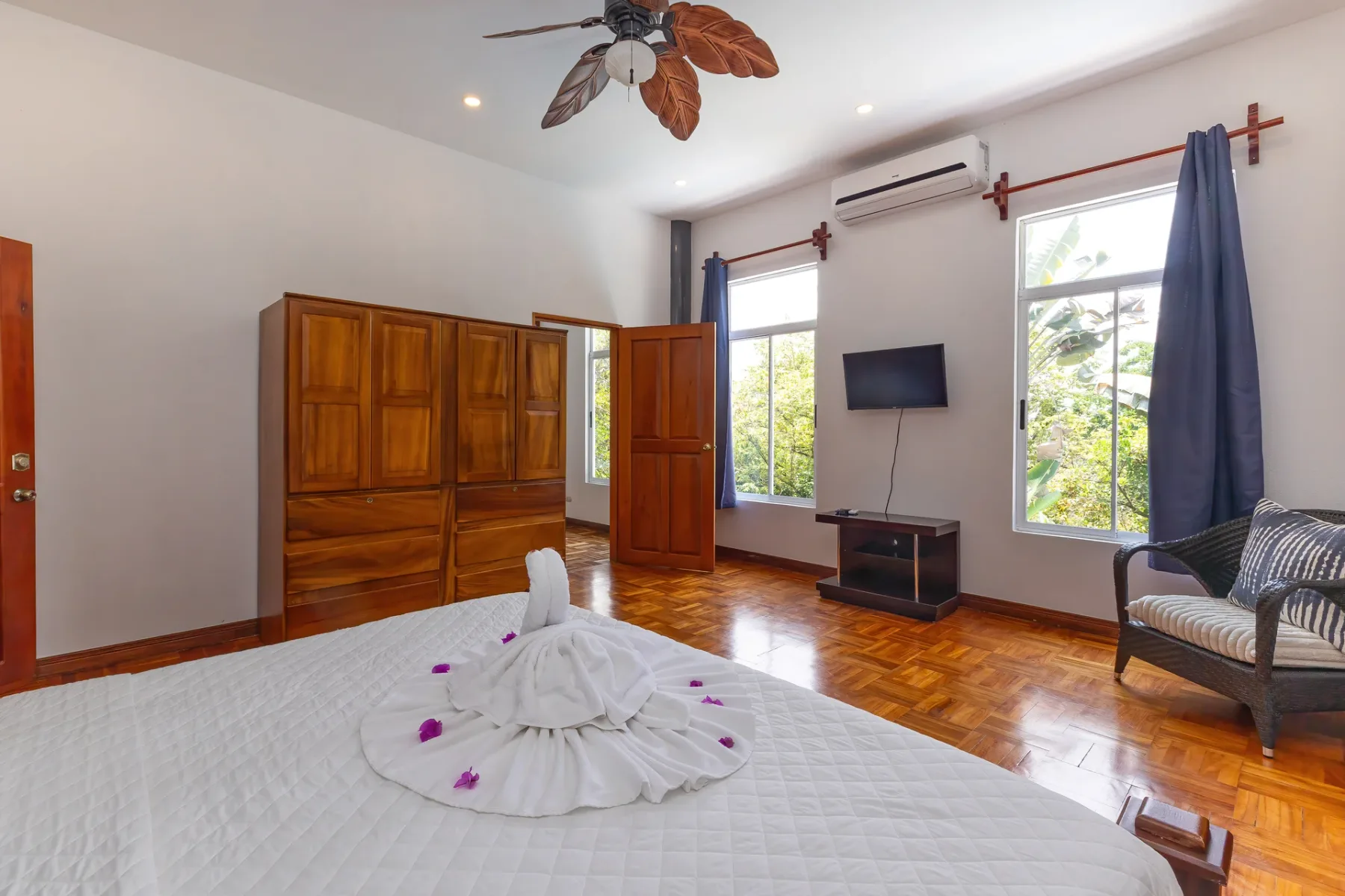 A bright bedroom with a large bed, wooden wardrobe, and windows overlooking greenery.