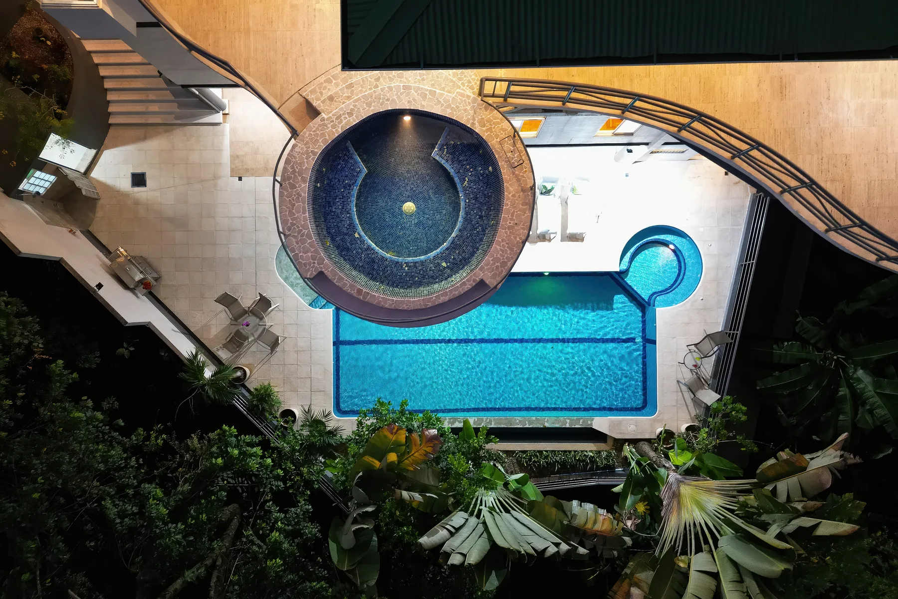 An outdoor swimming pool with a circular spa, surrounded by tropical greenery.