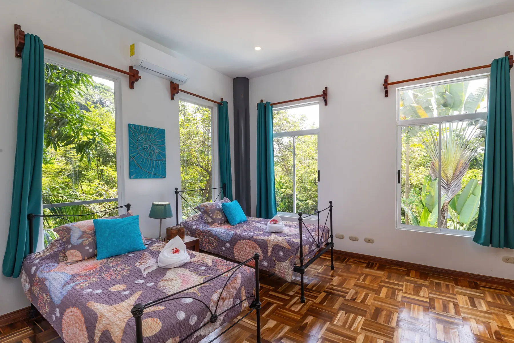 A bright bedroom with two beds, teal curtains, and windows overlooking greenery.