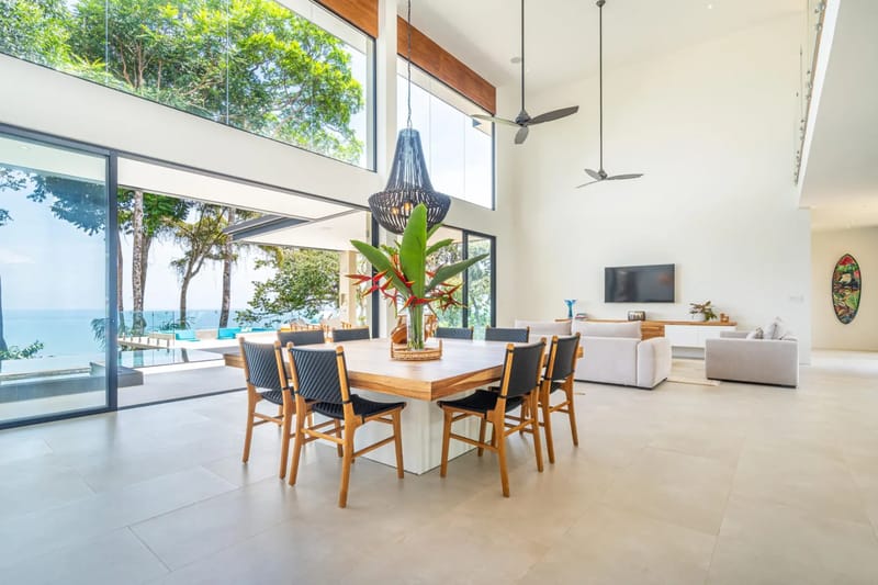 Casa La Vista · 15. The double-height dining area is filled with natural light from floor-to-ceiling glass doors and windows, offering seamless ocean views framed by tropical foliage.