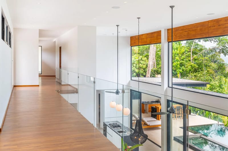 Casa La Vista · 8. This bright, open multi-level interior provides an upper walkway with glass railings, overlooking a modern kitchen below and offering views of lush tropical jungle through large windows.