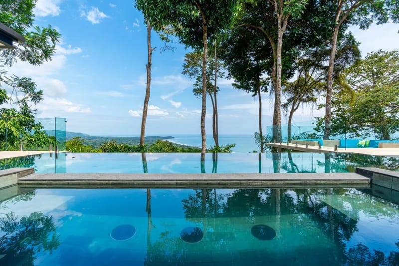 Casa La Vista · 18. An expansive infinity pool offers uninterrupted ocean and coastline views under a bright blue sky, framed by tropical trees and transparent glass railings.