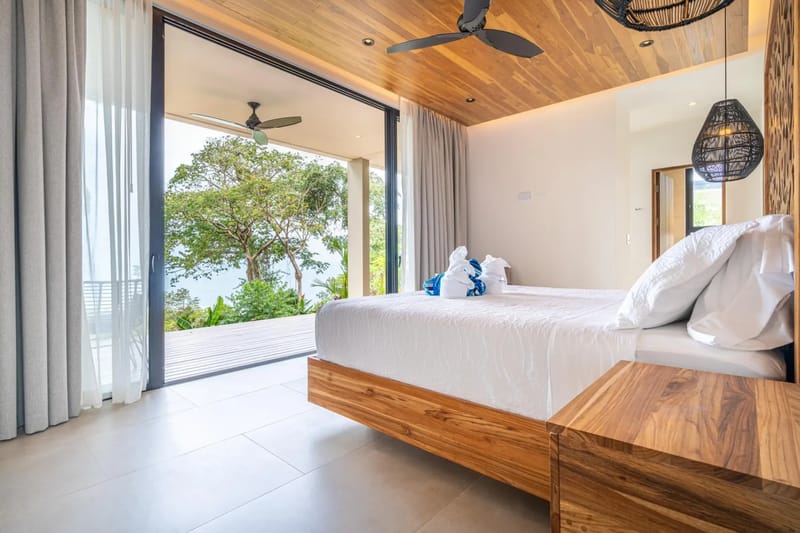 Casa La Vista · 19. This bedroom features a king-size bed, a wooden ceiling, and full-height glass doors opening to a private terrace with tropical foliage and ocean views, allowing abundant natural light.