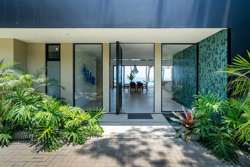 Casa La Vista · 9. The entrance, featuring an open pivot door and expansive glass, offers immediate views of the ocean through the bright interior living and dining spaces, surrounded by tropical landscaping.