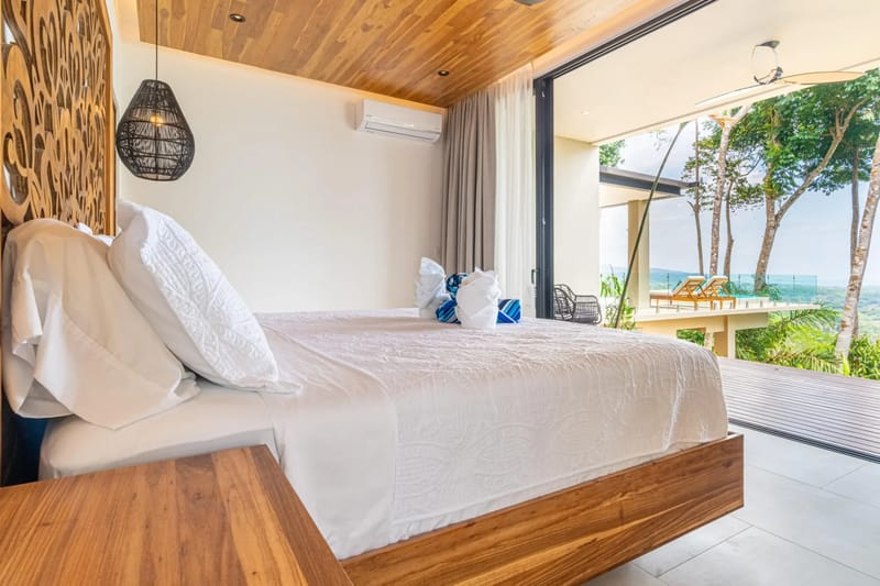 Casa La Vista · 14. A bedroom featuring a king bed with a carved wooden headboard and a wood-paneled ceiling, bathed in natural light from sliding glass doors that lead to a private terrace overlooking the ocean.