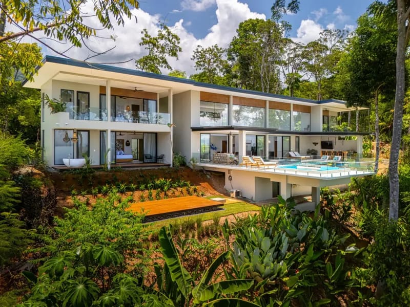 Casa La Vista · 16. The multi-level modern villa, featuring extensive glass and open-air terraces, is immersed in a vibrant tropical landscape, offering expansive outdoor living with an infinity pool and jacuzzi overlooking distant views.