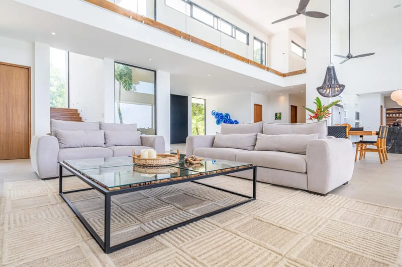 Casa La Vista · 10. This double-height living room features large windows with views of tropical greenery, abundant natural light, and an open connection to the dining and kitchen areas, overlooked by a second-story walkway.