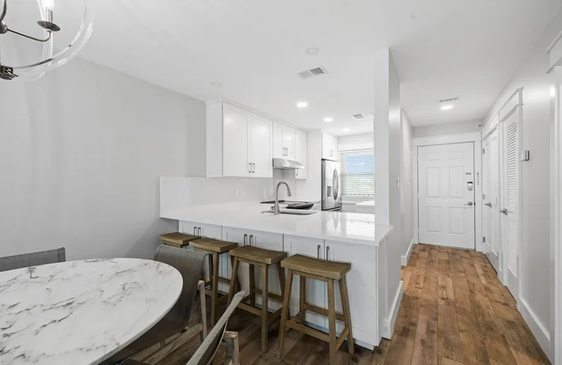 A bright kitchen with a counter and stools opens onto a wood-floored hallway.