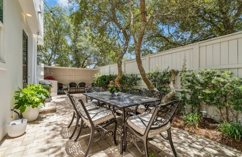 An outdoor patio with a dining table and seating beneath leafy trees.