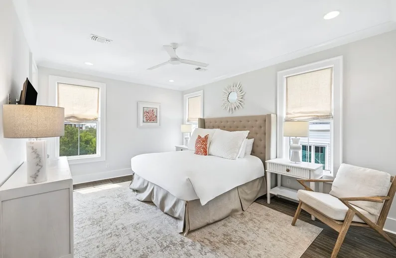 Un dormitorio luminoso con una cama grande, dos ventanas y una silla acolchada.
