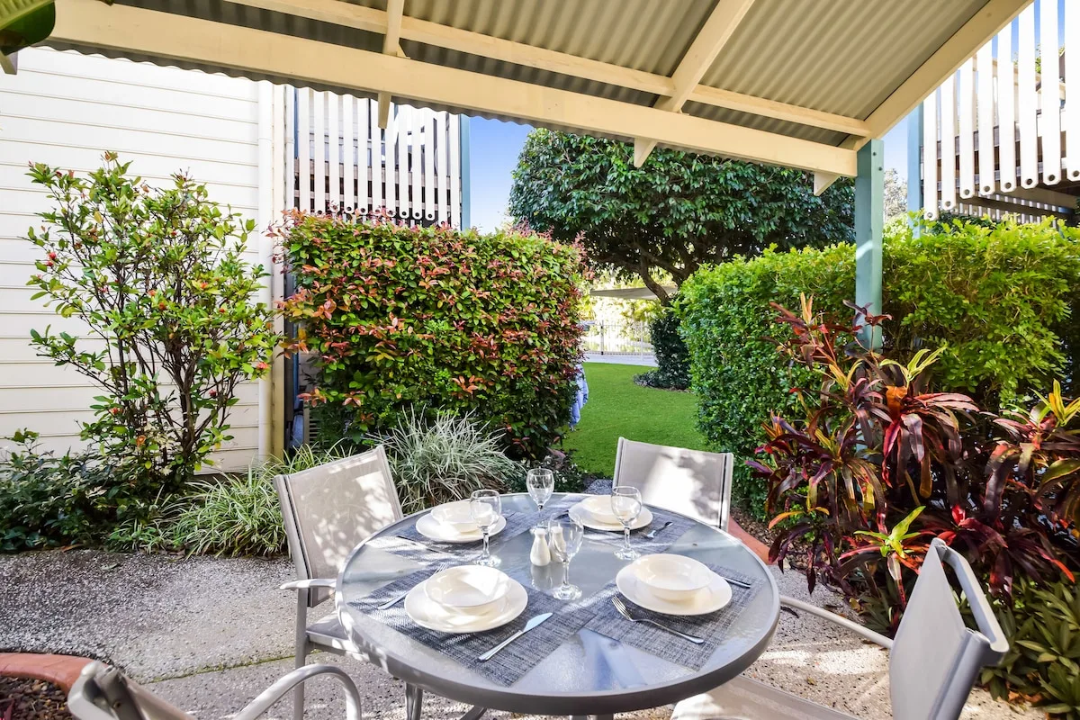 A covered outdoor dining area with a round table set among leafy garden plants.