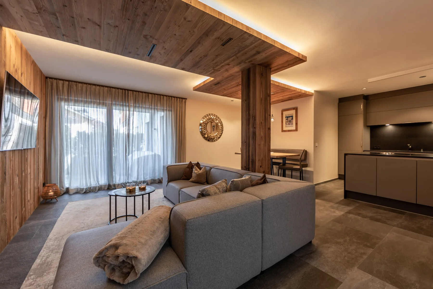 A modern living room with gray seating, large windows, and an open kitchen.