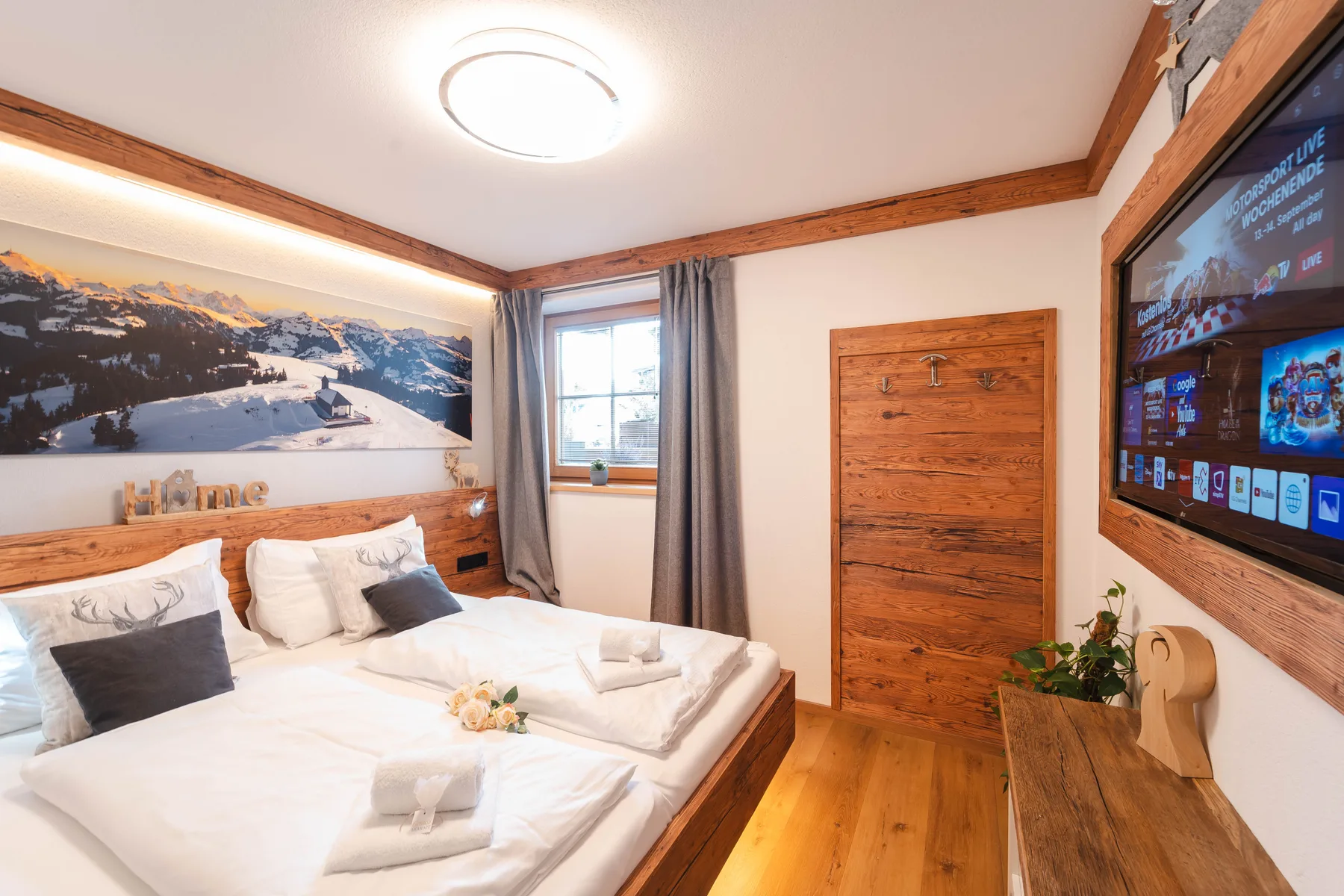 A bedroom with a wooden bed, mountain photograph, and wall-mounted television.